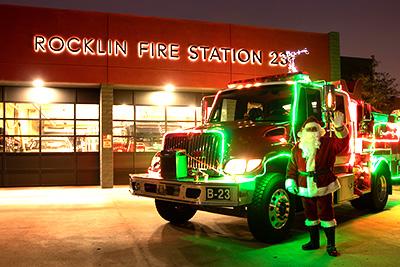Santa waving and standing next to a fire truck lit up with Christmas lights in front of Rocklin Fire Station 23
