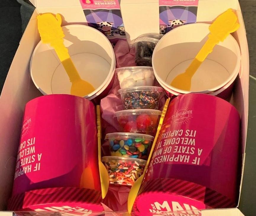 Two 32-oz frozen yogurt containers, five toppings, four cups and spoons, and two bookmarks are nestled in a cardboard carrying box.