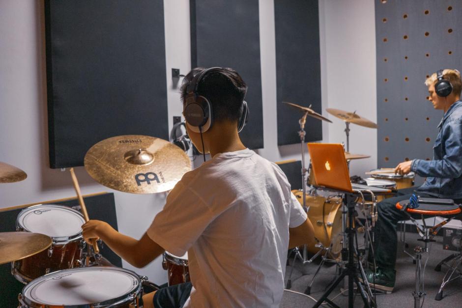 Two drummers practice on their own sets in a spacious music room.