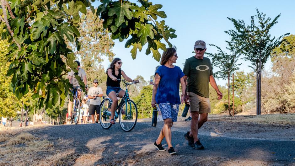 A couple walks on a path at the Sunset Whitney Recreation Area. A cyclist on a beach cruiser is also seen in the background. The photo is framed by branches from a developed tree on the left and a young sapling on the right. 
