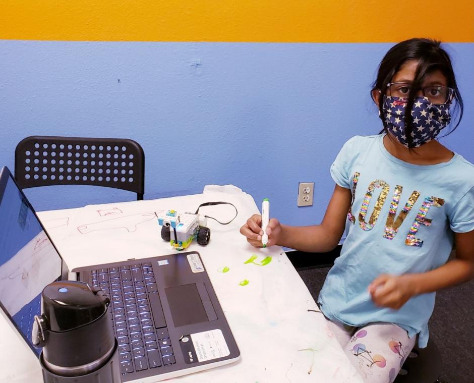 A young girl sits at a desk with a laptop and robot nearby.