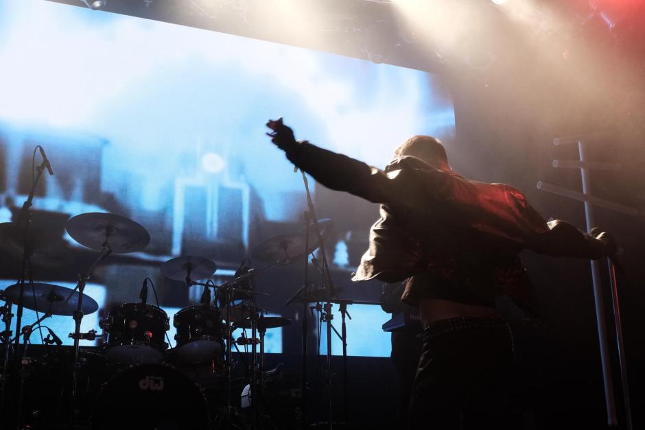 Man in the right side of the photo with back to the audience. Arms spread out wide. Digital screen behind the singer is blue with a blurred image on it. This is a live performance photo with stage lights. 