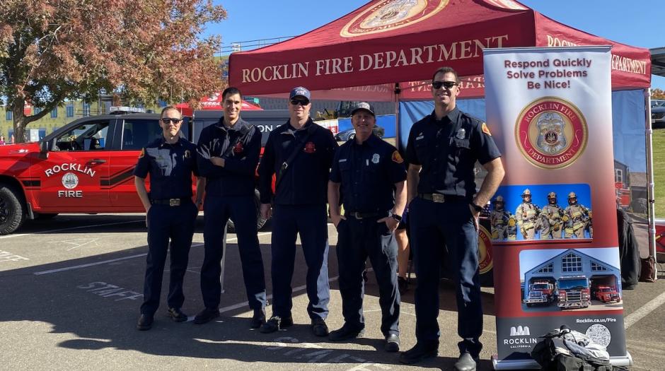 Five members of the Rocklin Fire Department standing and smiling in front of a pop-up recruitment tent.