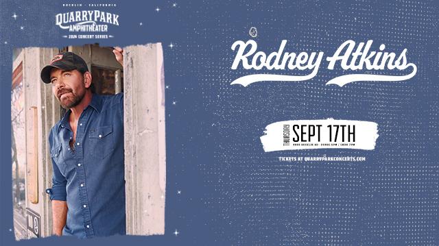 Rodney Atkins concert poster for Quarry Park Amphitheatre in Rocklin, California featuring Rodney Atkins wearing a blue denim shirt and camouflage baseball cap, leaning against a weathered white doorframe with an American flag visible in the background. Top text reads "Quarry Park Amphitheater 2026 Concert Series" in white. Artist name "Rodney Atkins" appears in black script lettering with white outline across the top of the photo. Bottom section states "Thursday Sept 17th, 4000 Rocklin Rd, Doors 6PM / Show 7PM" on a cream brushstroke banner. Footer reads "Tickets at quarryparkconcerts.com" in white text against a dark blue starry background.